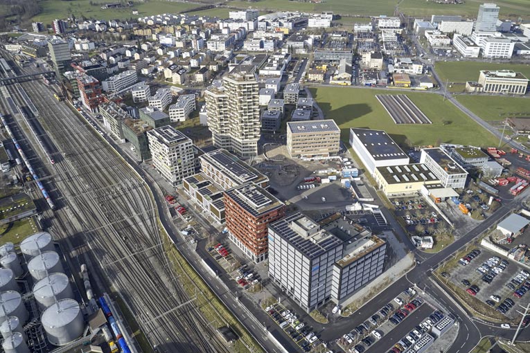 Bilan carbone : Le quartier de Suurstoffi à Risch-Rotkreuz s’étend sur 10 ha sur une friche industrielle en bordure de la voie ferrée. Au premier plan, Suurstoffi 22, pionnier des tours à structure bois de moyenne hauteur en Suisse. Et au tout premier plan, en marge du quartier, les groupes de refroidissement du réseau de chaleur. [©Zug Estates]